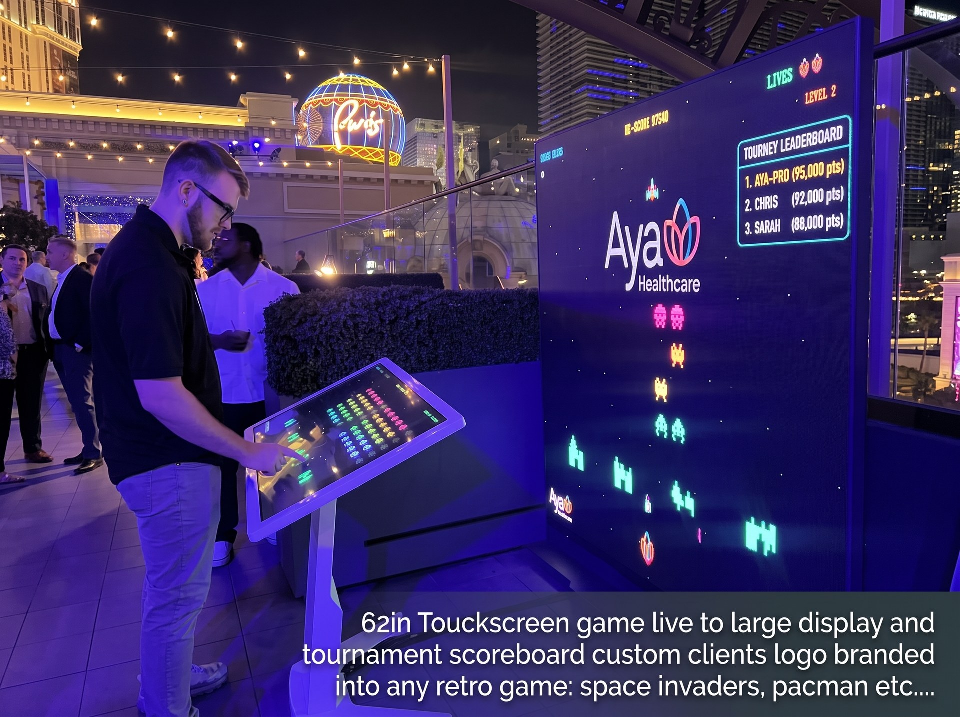Guest playing a custom-branded retro game on a 62-inch touch screen kiosk with a giant tournament scoreboard at a Las Vegas event